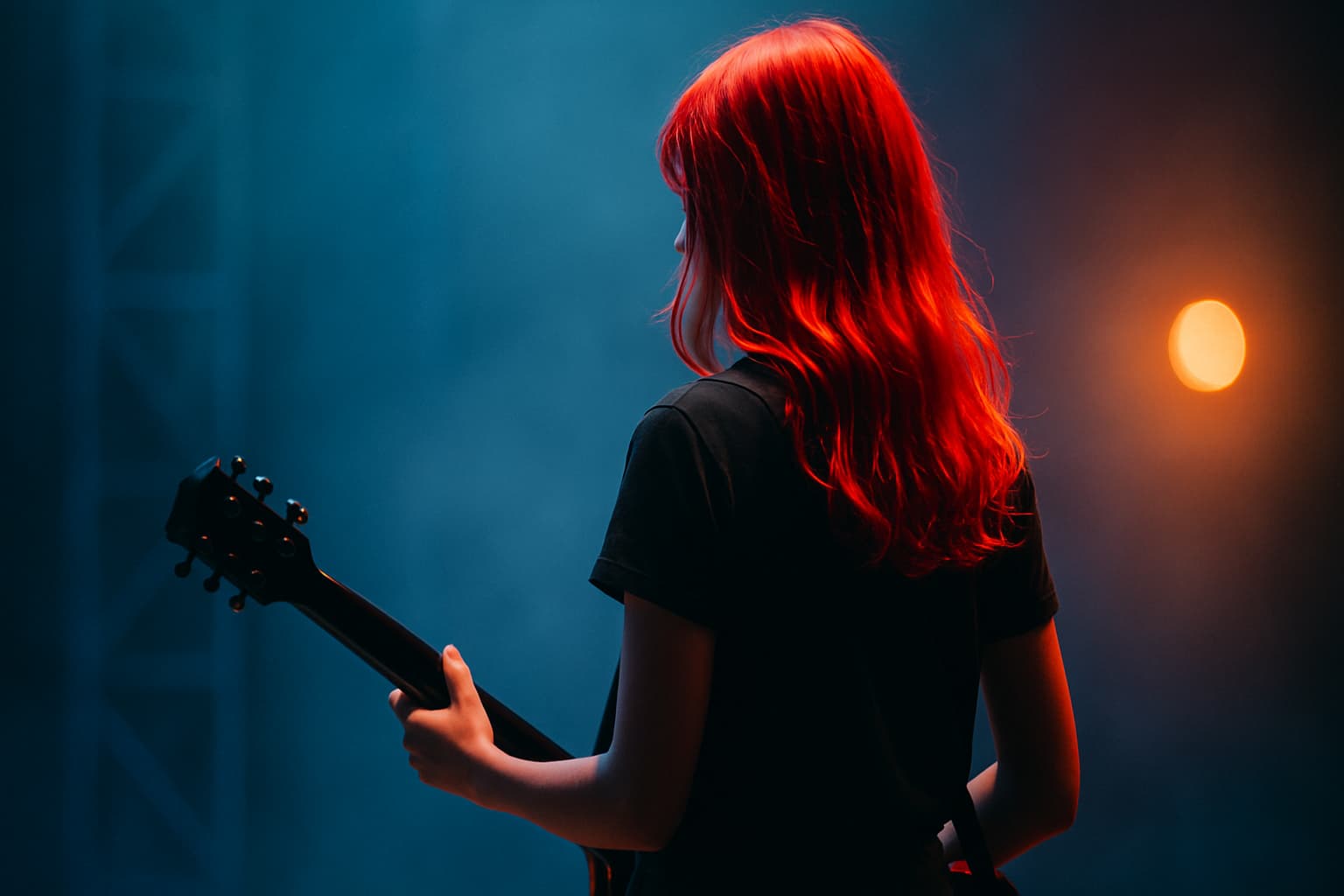 ステージ袖に立つ赤髪の女性が黒いエレキギターを持つ。青とオレンジのライトに照らされた後ろ姿のシネマ風写真。